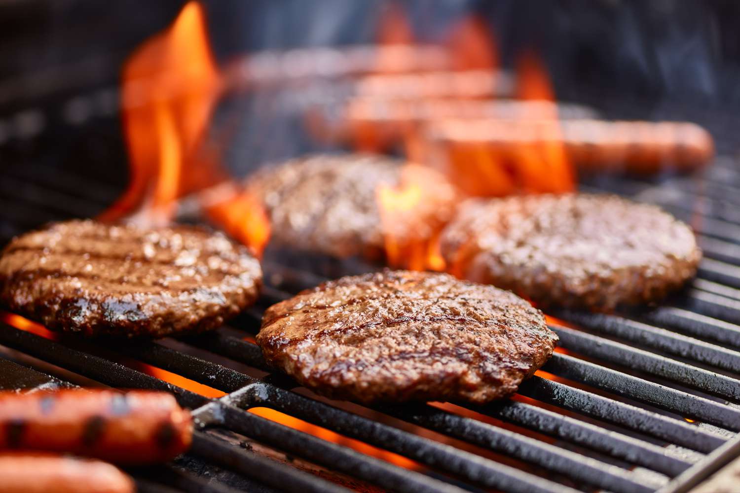 Hamburger patties and hot dogs cooking on a grill with flames