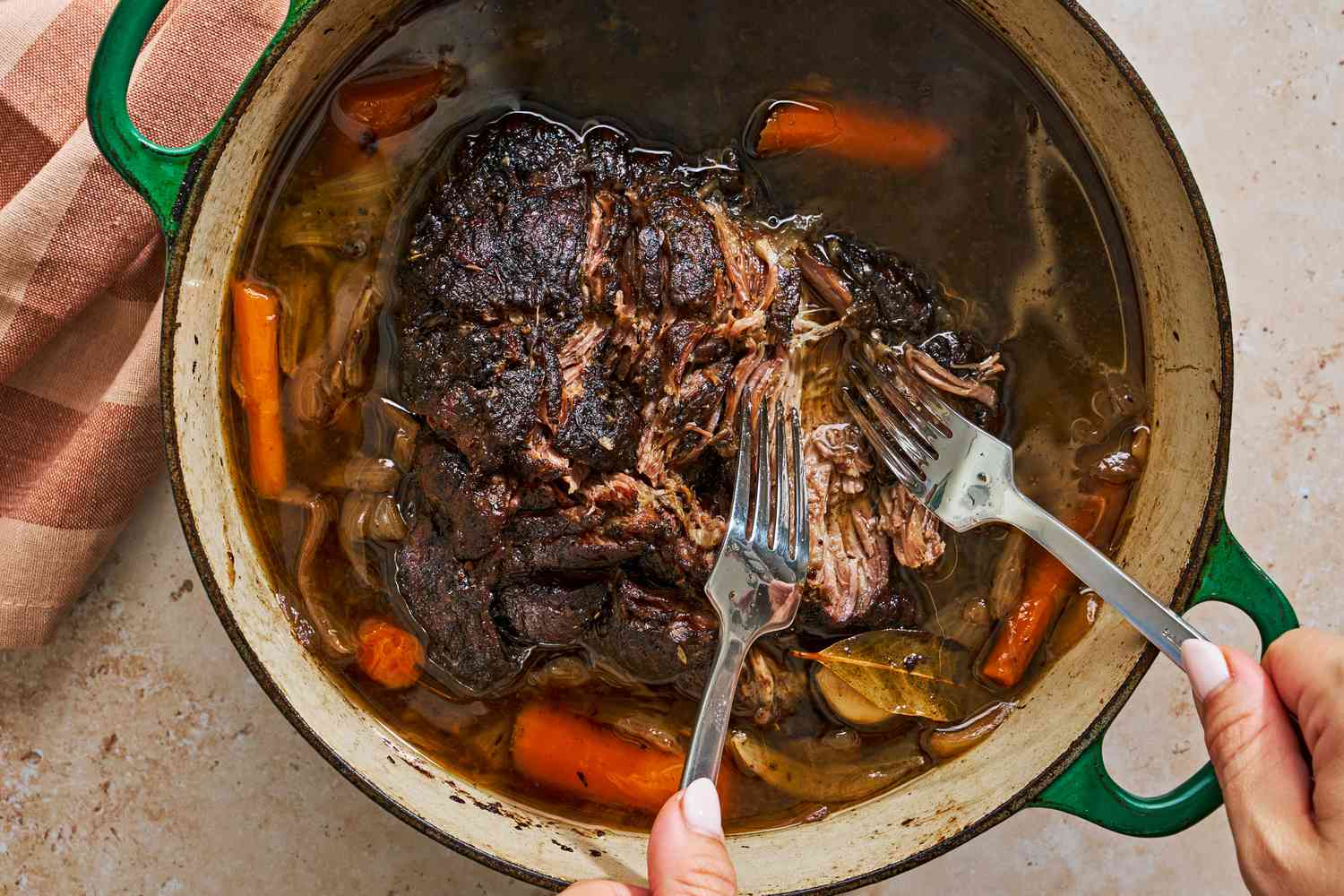 A Dutch oven of pot roast and hands holding two large forks, flaking apart the meat