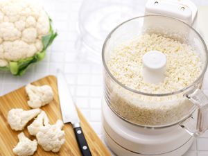 lidless food processor with processed cauliflower in it, head of cauliflower on the side and a few cauliflower florets on a cutting board with a knife