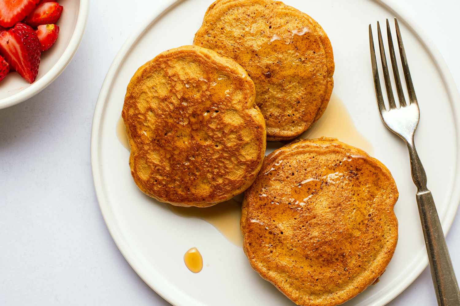 A plate with almond butter pancakes and a fork a bowl with strawberries on the side