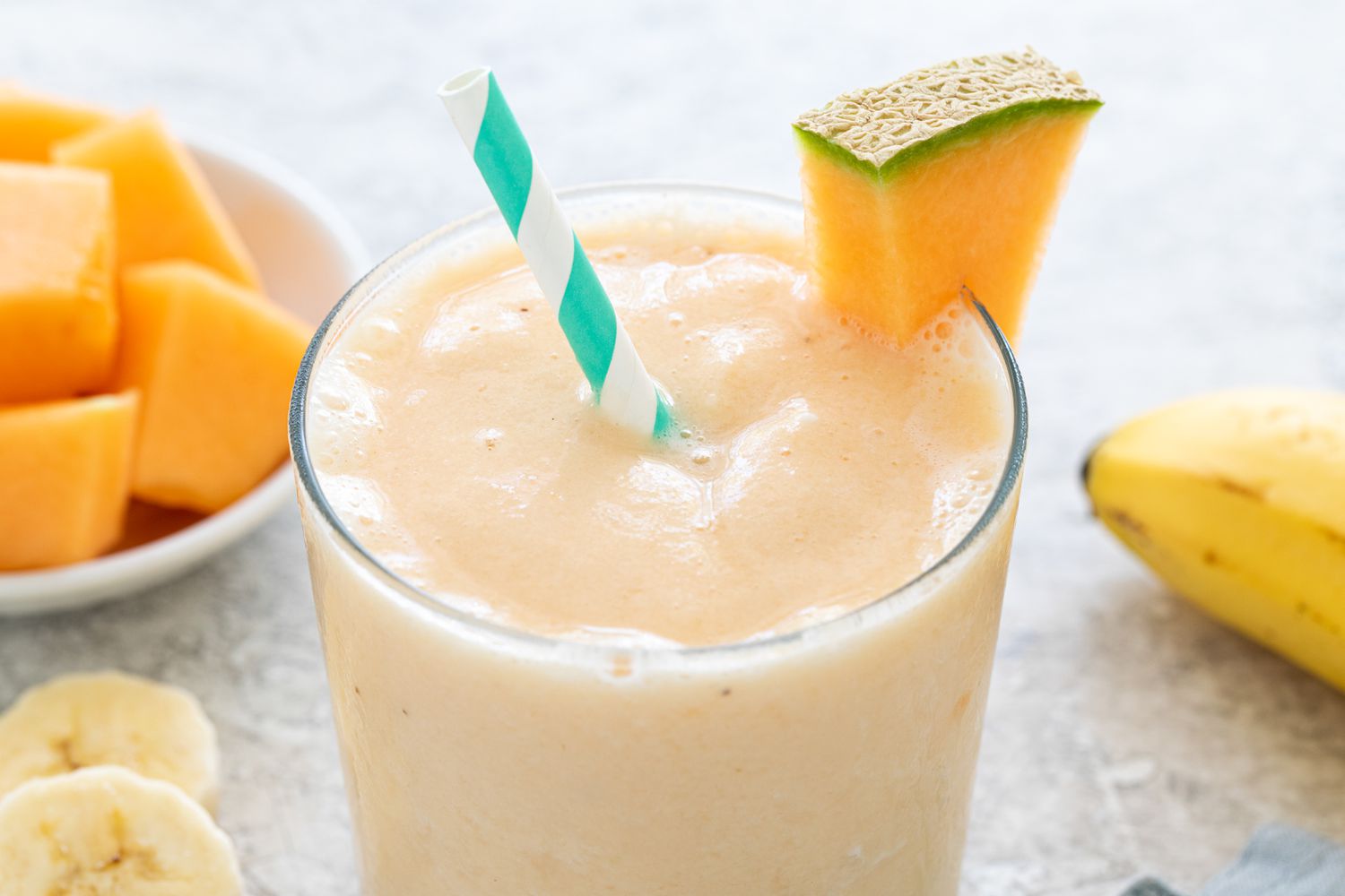 Glassful of Cantaloupe Smoothie With a Slice of Cantaloupe on the Glass Rim, and in the Surroundings, a Bowl of Cantaloupe and Slices of Bananas and a Whole Banana on the Counter
