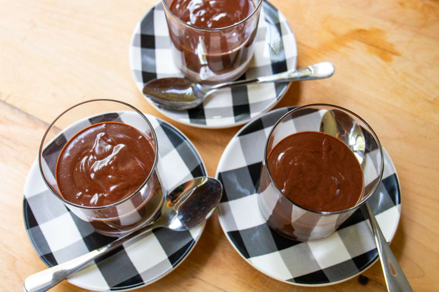 Three servings of chocolate dessert in glasses on checkered plates with spoons next to them
