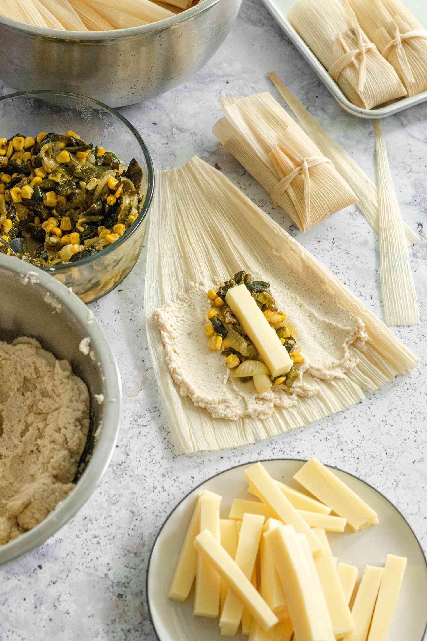 Poblano, Corn, and Cheese Added to Masa on a Corn Husk and Surrounded by a Bowl of Poblano and Corn Mix, Bowl of Masa, and Plate of Cheese Strips Tamales 