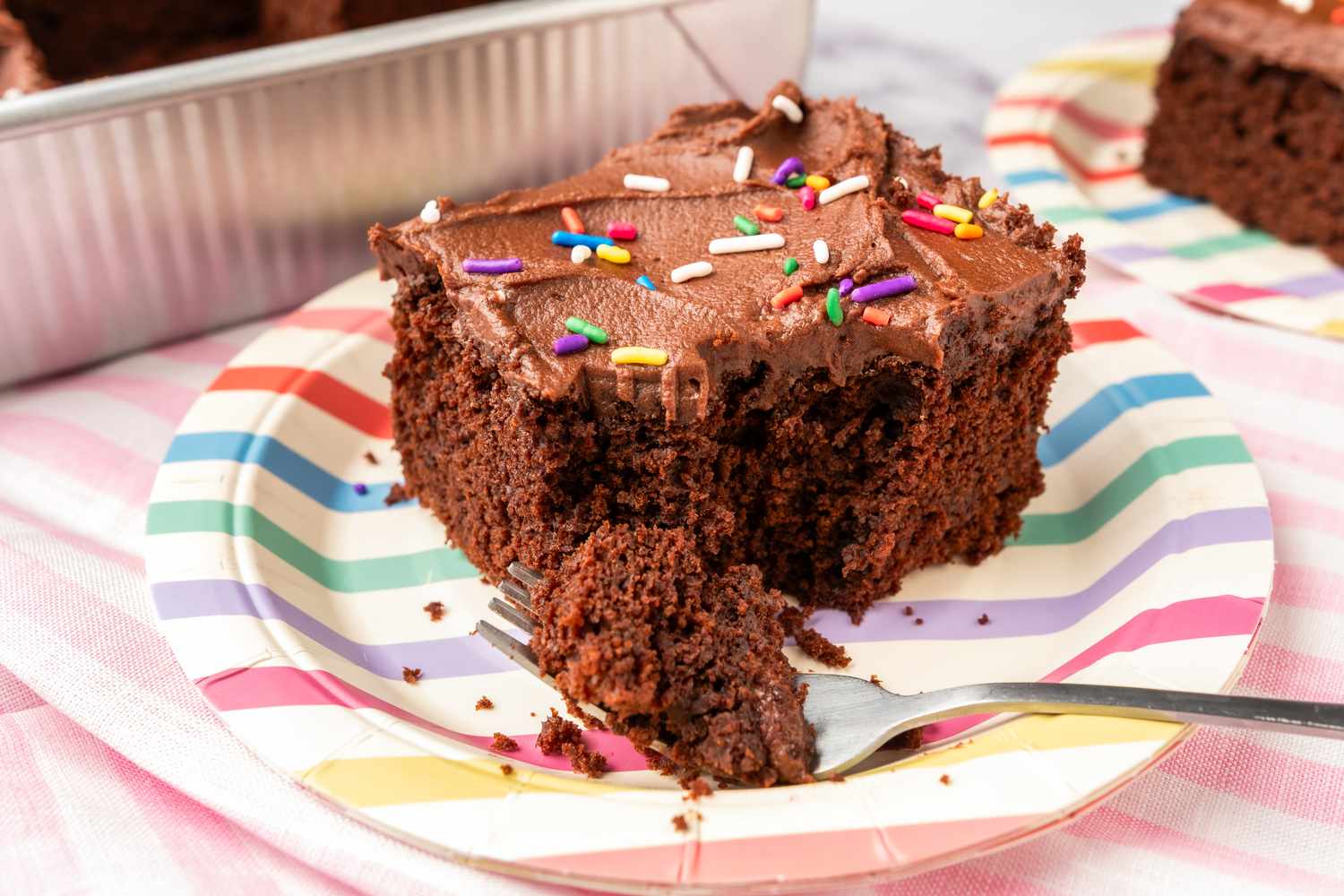 Slice of chocolate cake with frosting and sprinkles on a striped plate fork with a bitesized piece