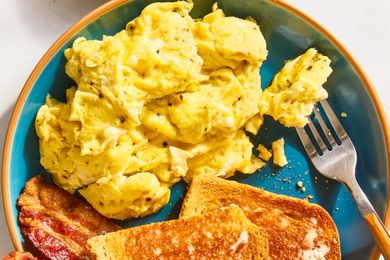 Scrambled eggs served on a plate with toast and bacon a fork placed to the side