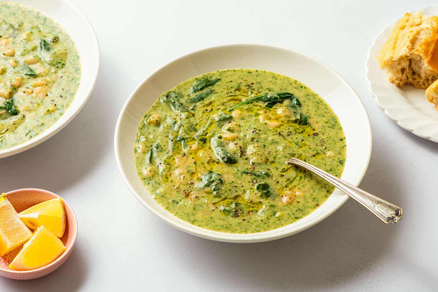Angled view of a white bowl of white bean and spinach soup with a spoon next to a plate of bread and bowl of lemon slices