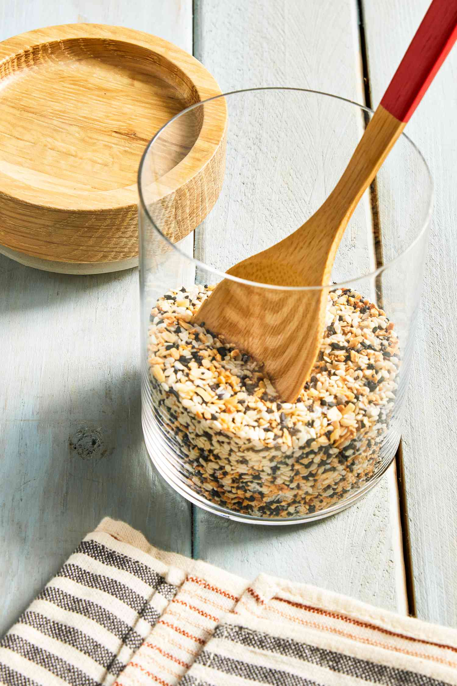 Everything Bagel Seasoning in a Tall Cylindrical Glass Jar With a Wooden Spoon In It, Next to a Wooden Circular Top and a Black and White Stripped Kitchen Towel