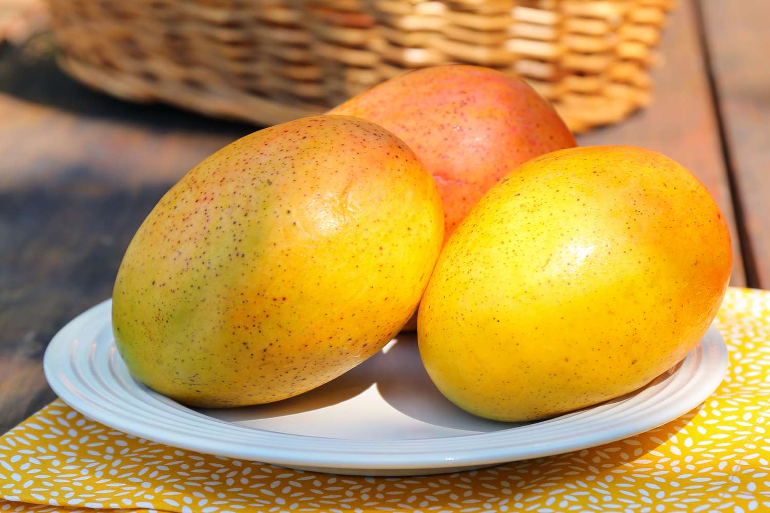 Three ripe mangos arranged on a white plate with a wicker basket in the background
