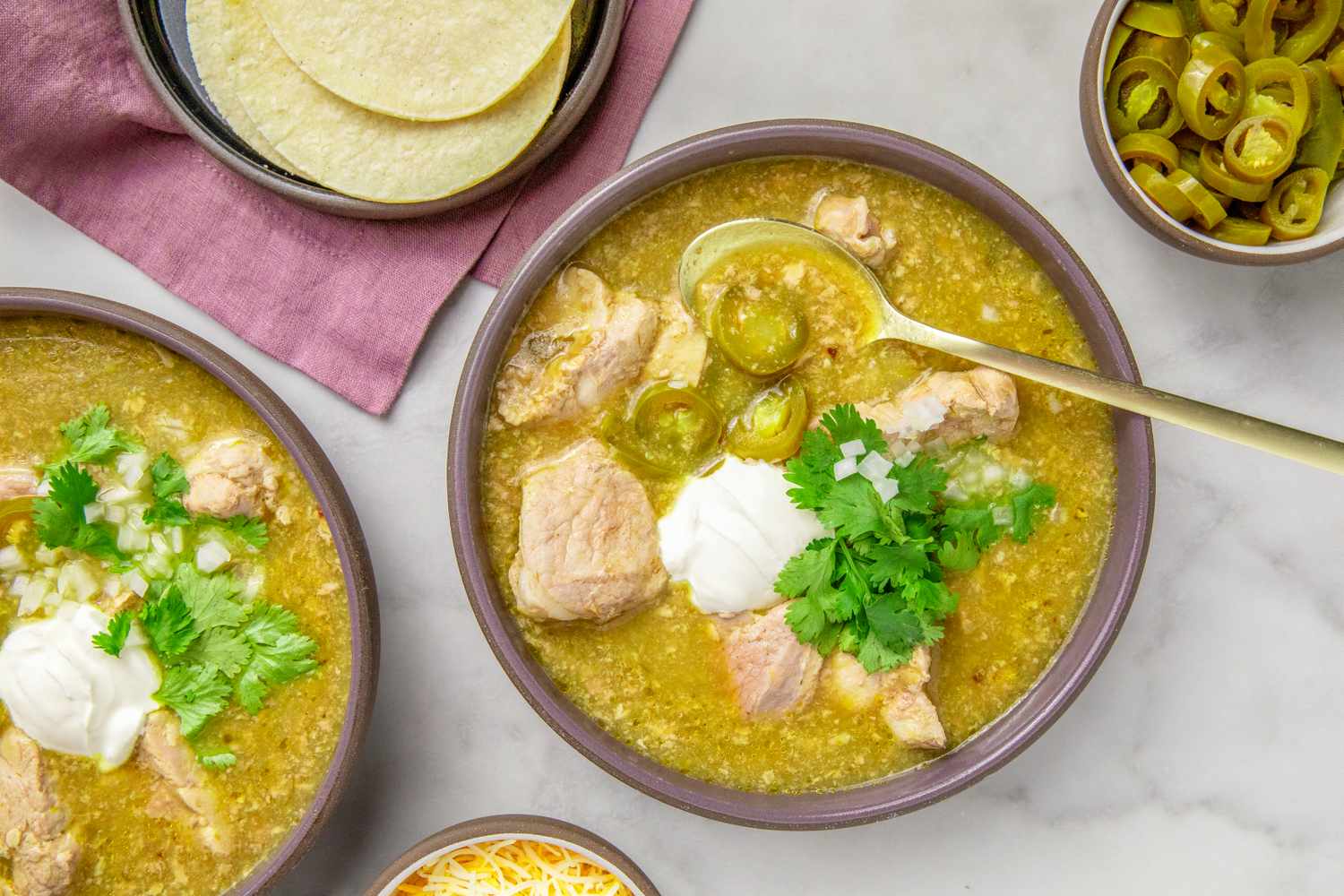Bowls of pork chile verde garnished with sour cream cilantro and jalapeo slices surrounded by tortillas and toppings