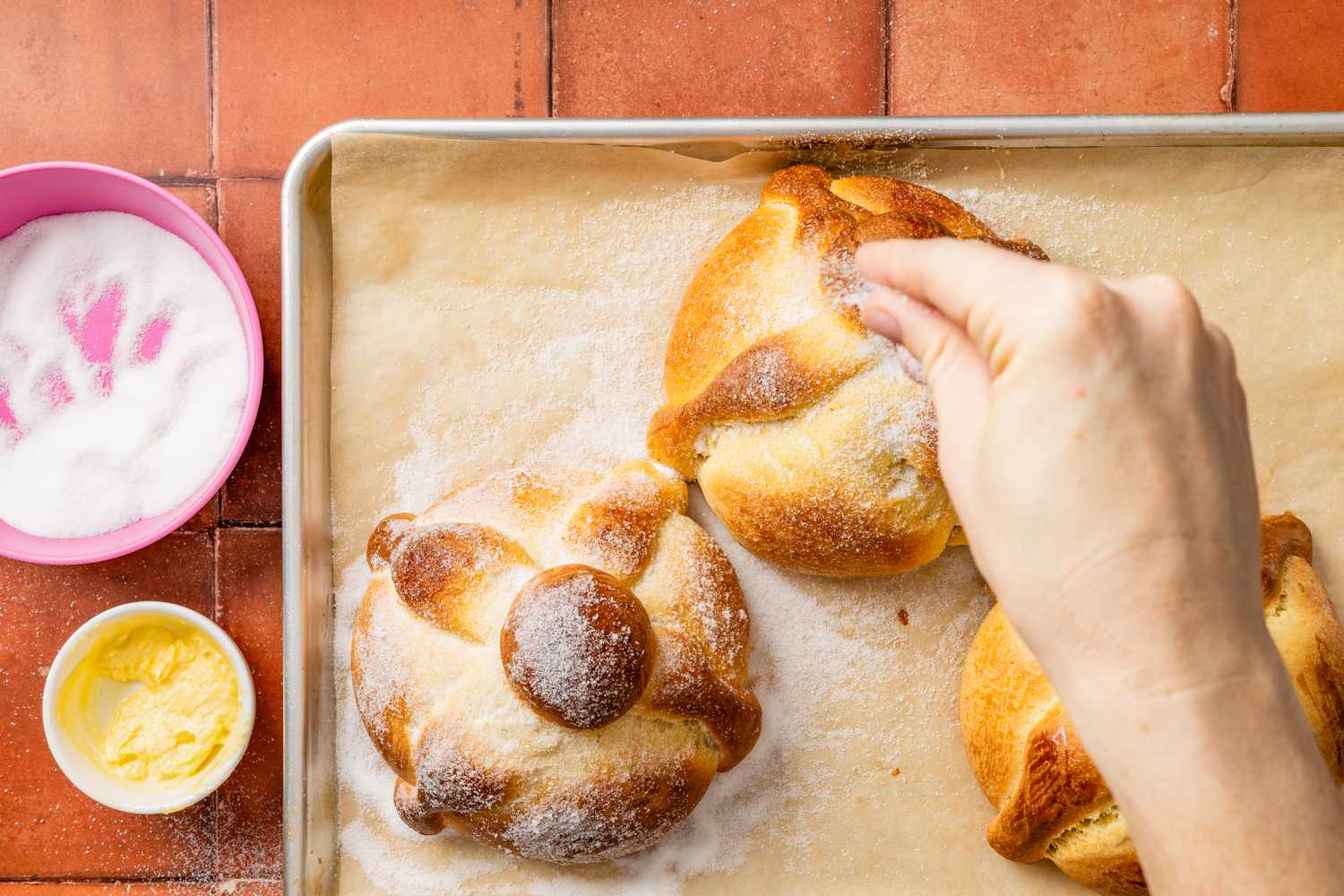 Pan de muerto sprinkled with sugar next to one already covered in sugar (all on a parchment paper lined baking sheet)