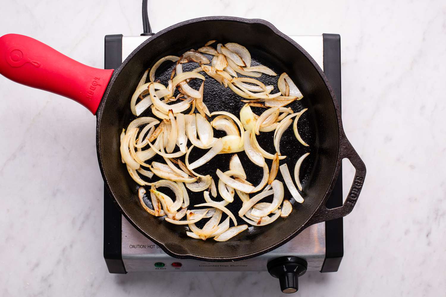 Overhead view of a cast iron skillet sauteing onions for Melting Cabbage recipe on a hot plate on a marble countertop