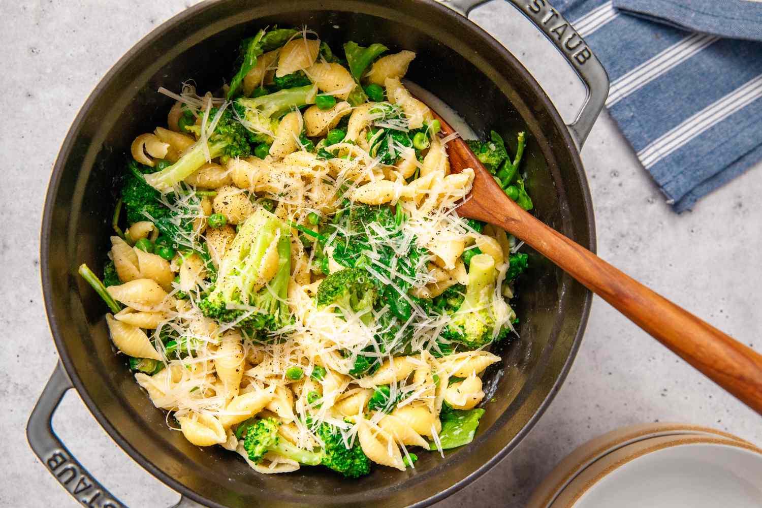 A pot of pasta shells mixed with vegetables and topped with grated cheese, served with a wooden spoon