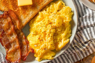 Photo of a plate of scrambled eggs, butter on toast, and slices of bacon on a white and gray kitchen towel with yellow lined illustration in the corner
