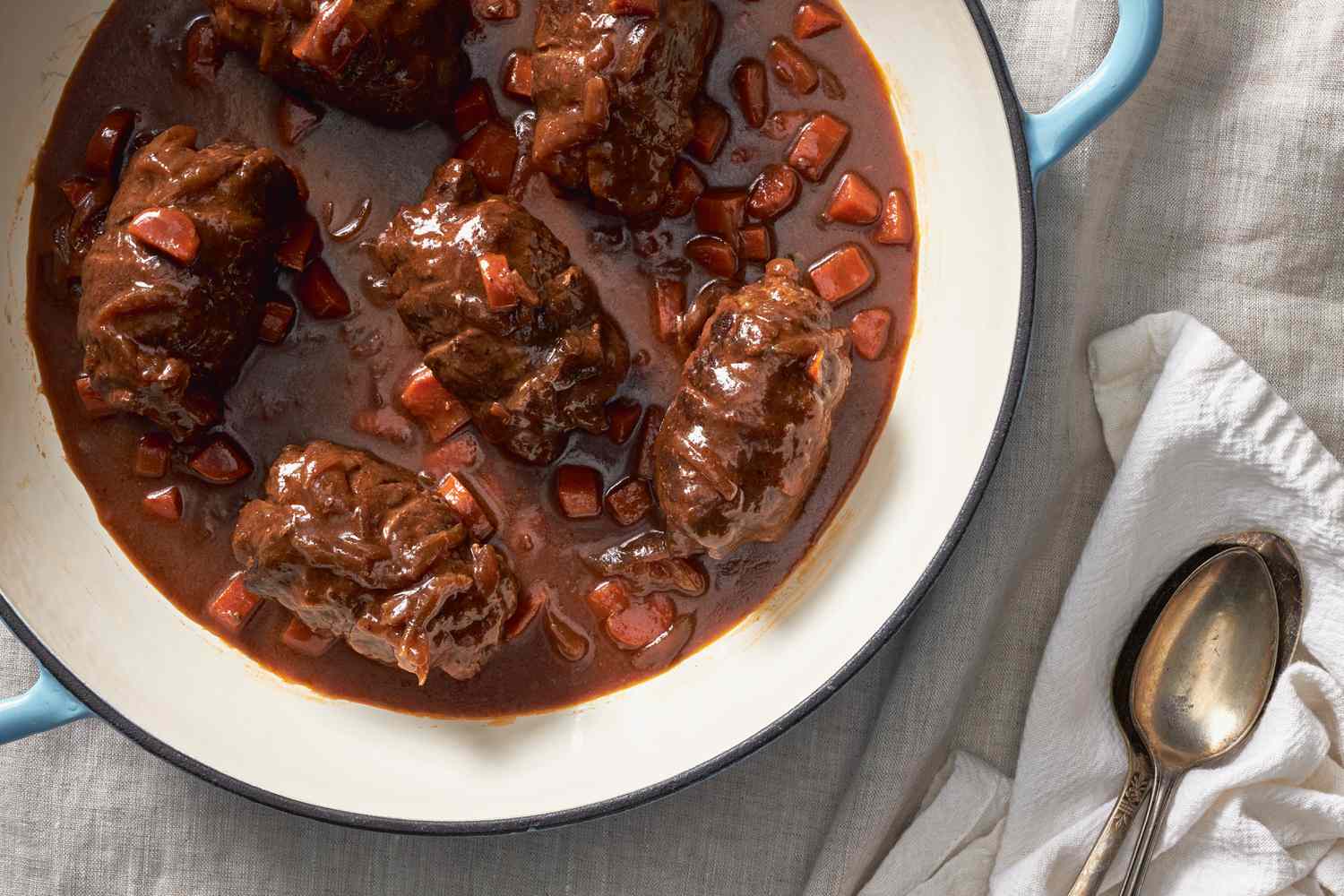 Rouladen in a Dutch oven next to some spoons on a table napkin