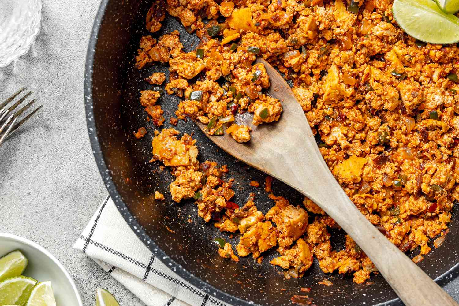 Copycat Sofritas in a Pan with a Wooden Spoon and Lime Wedges, Surrounded by a Bowl of Lime Wedges, a Glass of Water, and a Couple of Forks