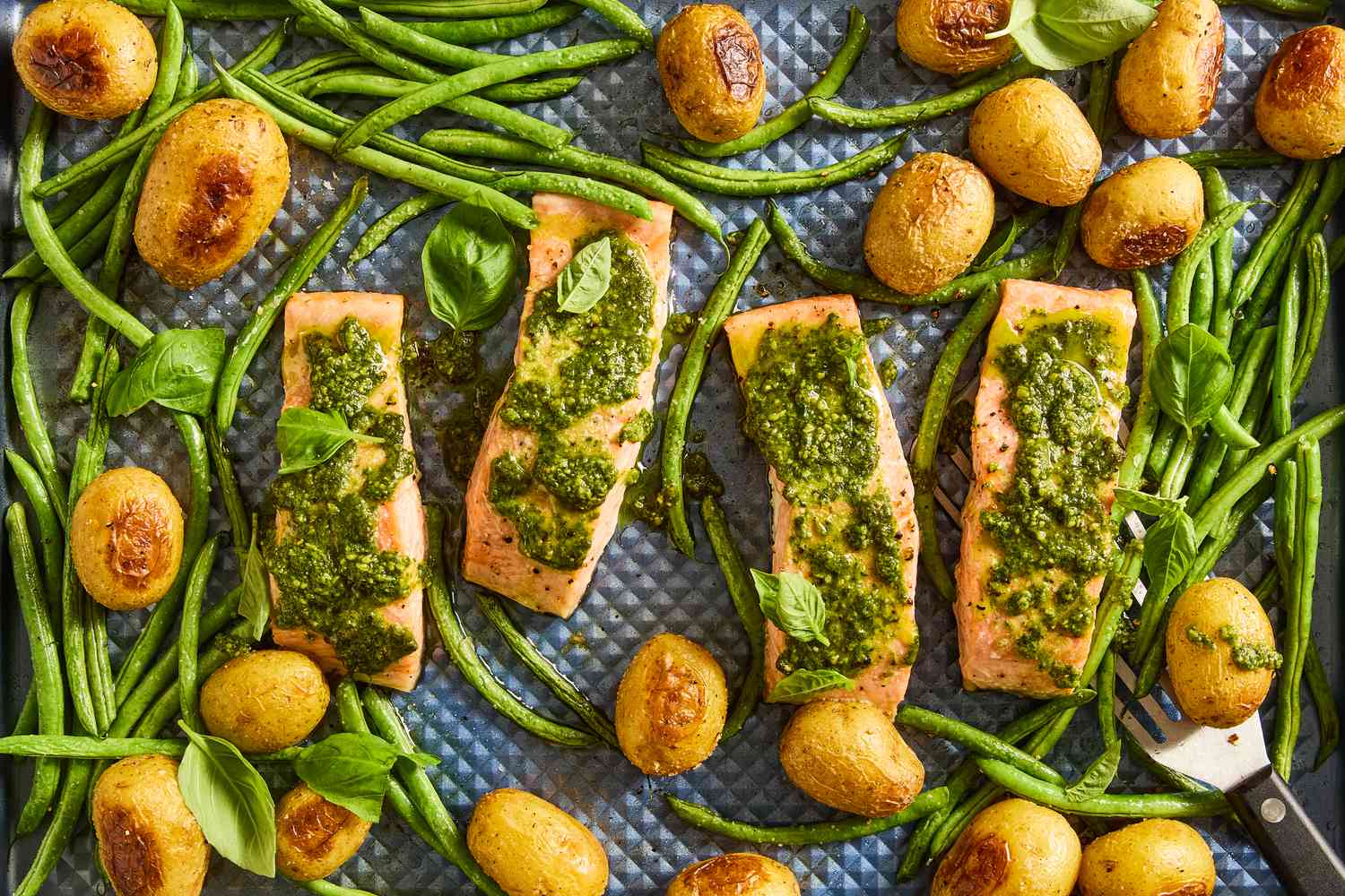 A sheetpan with salmon fillets topped with pesto, green beans, potatoes and basil