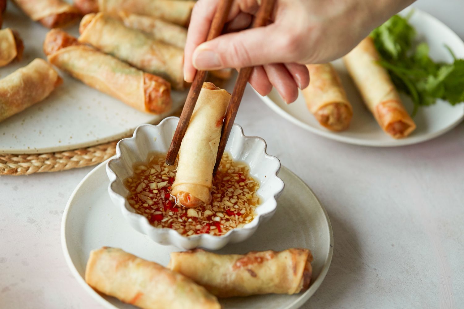 Dipping a spring roll into a sauce with more spring rolls on the plate and on a platter behind it.