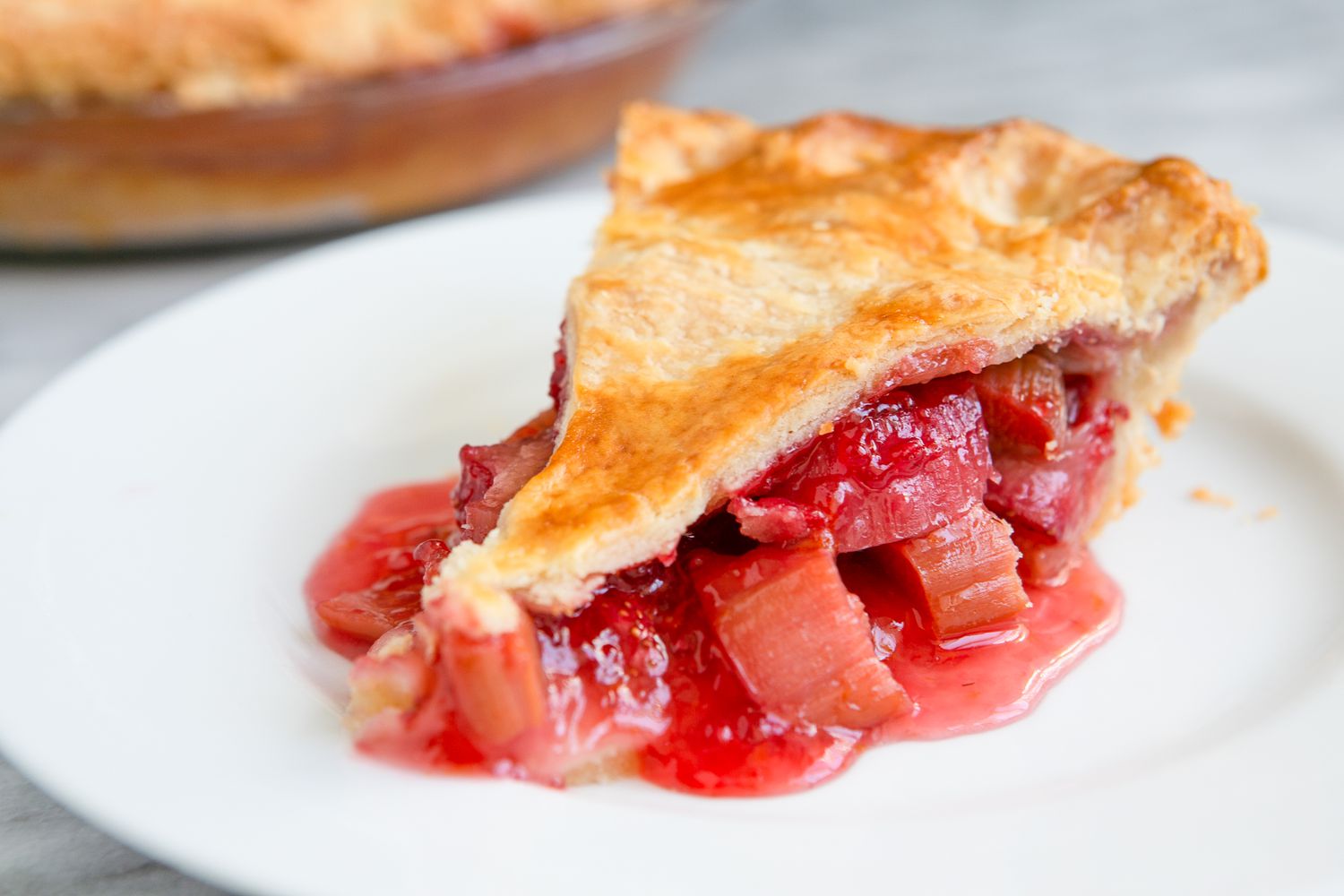 Slice of Strawberry Rhubarb Pie on a Plate, and in the Background, More in a Glass Pie Pan