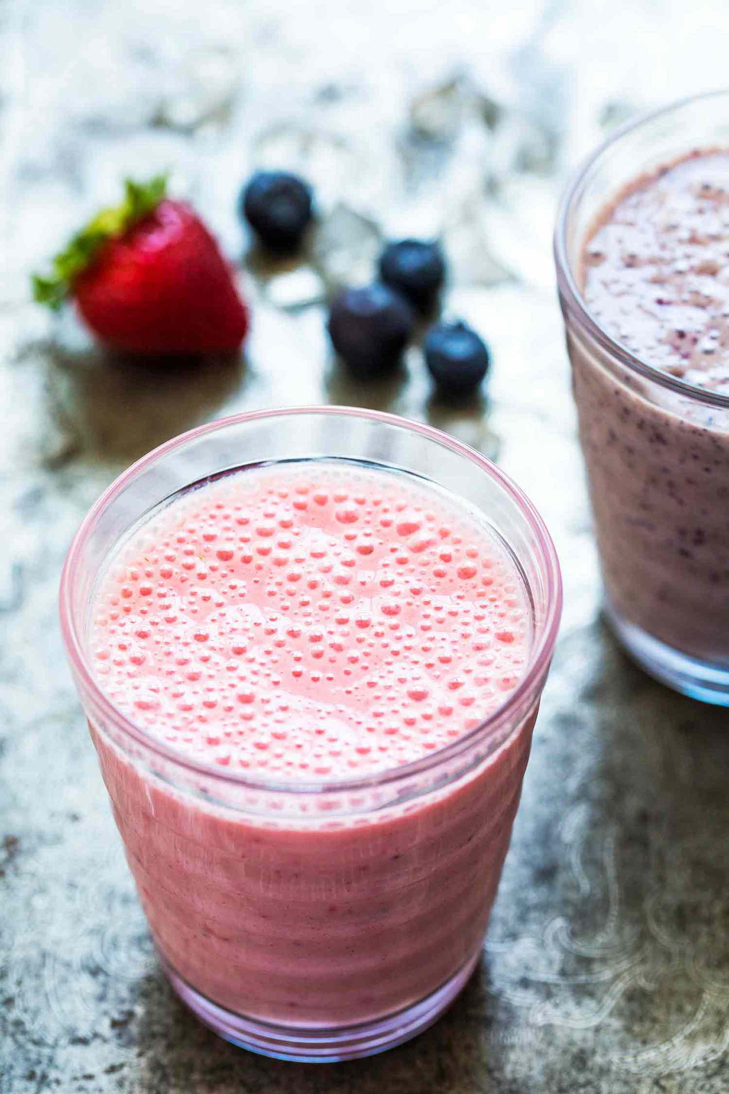Two glasses of berry and banana smoothies on a table surrounded by strawberries and blueberries