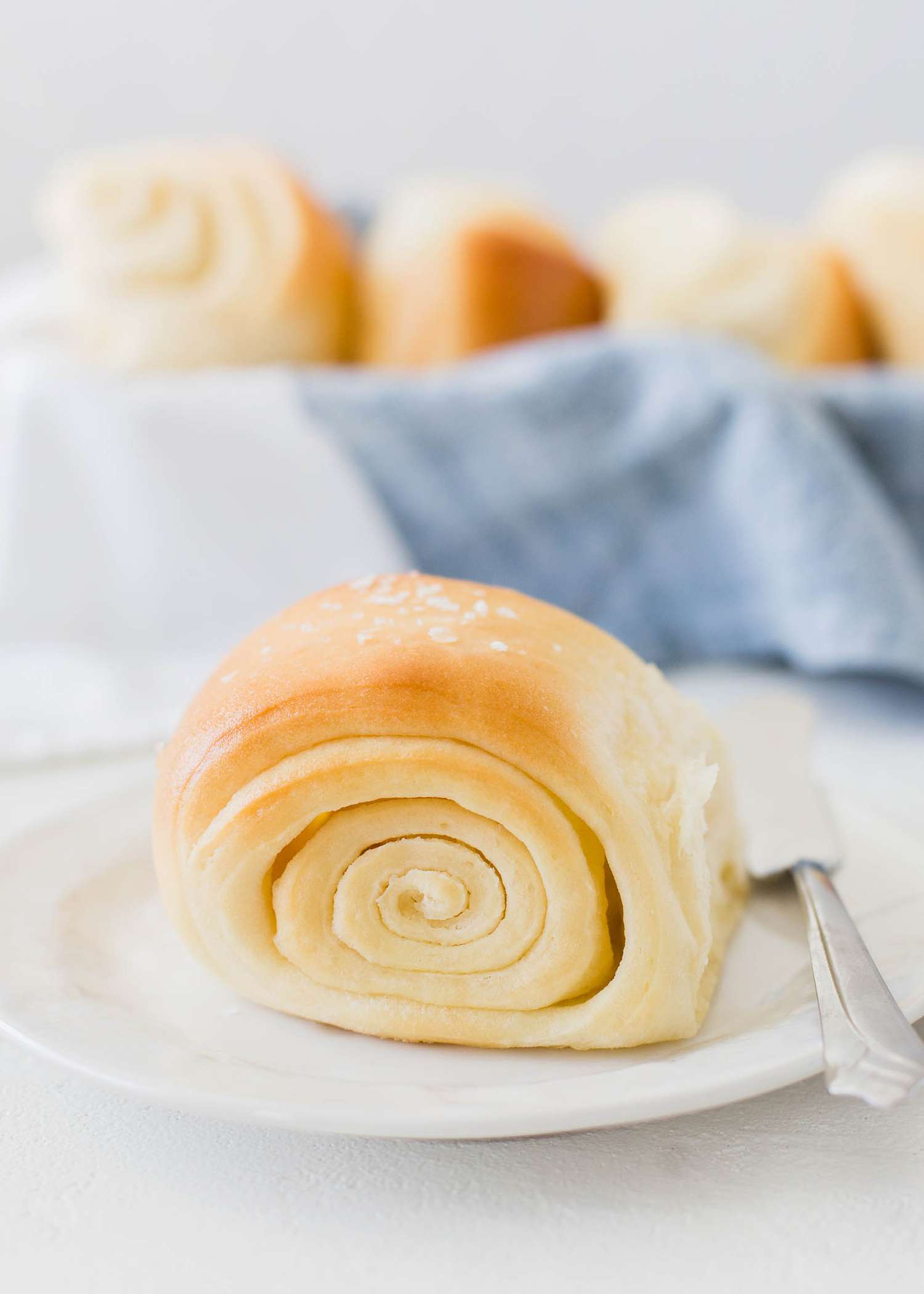 Lion House Roll on a plate with its swirls facing out. A basket of additional rolls is behind the plate.