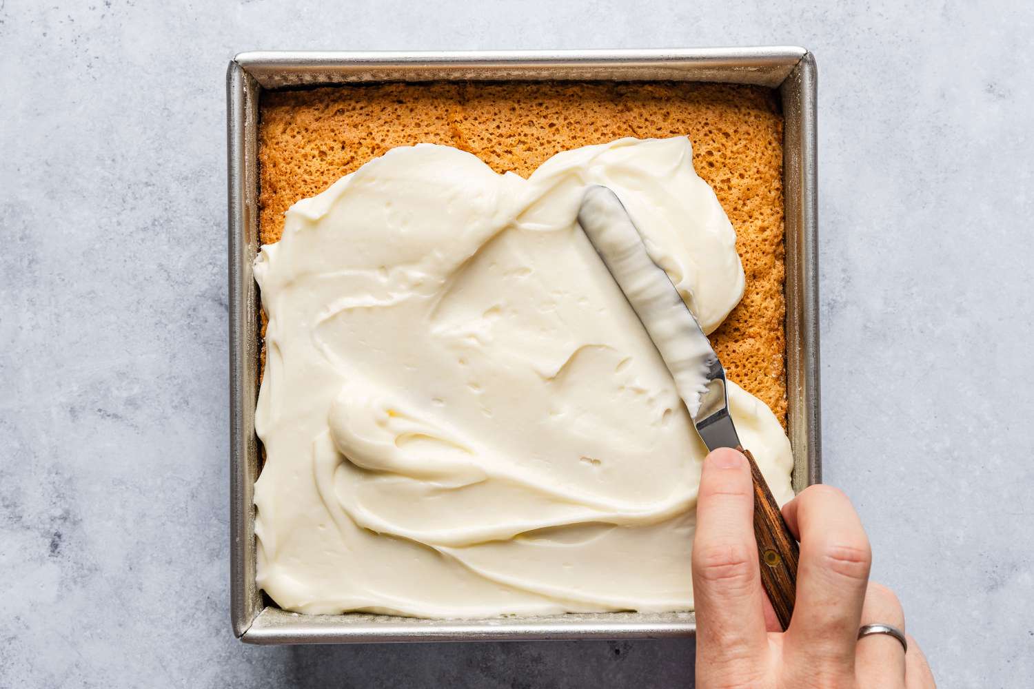 square tin with cooked cake, hand spreading icing on top - Feather Cake
