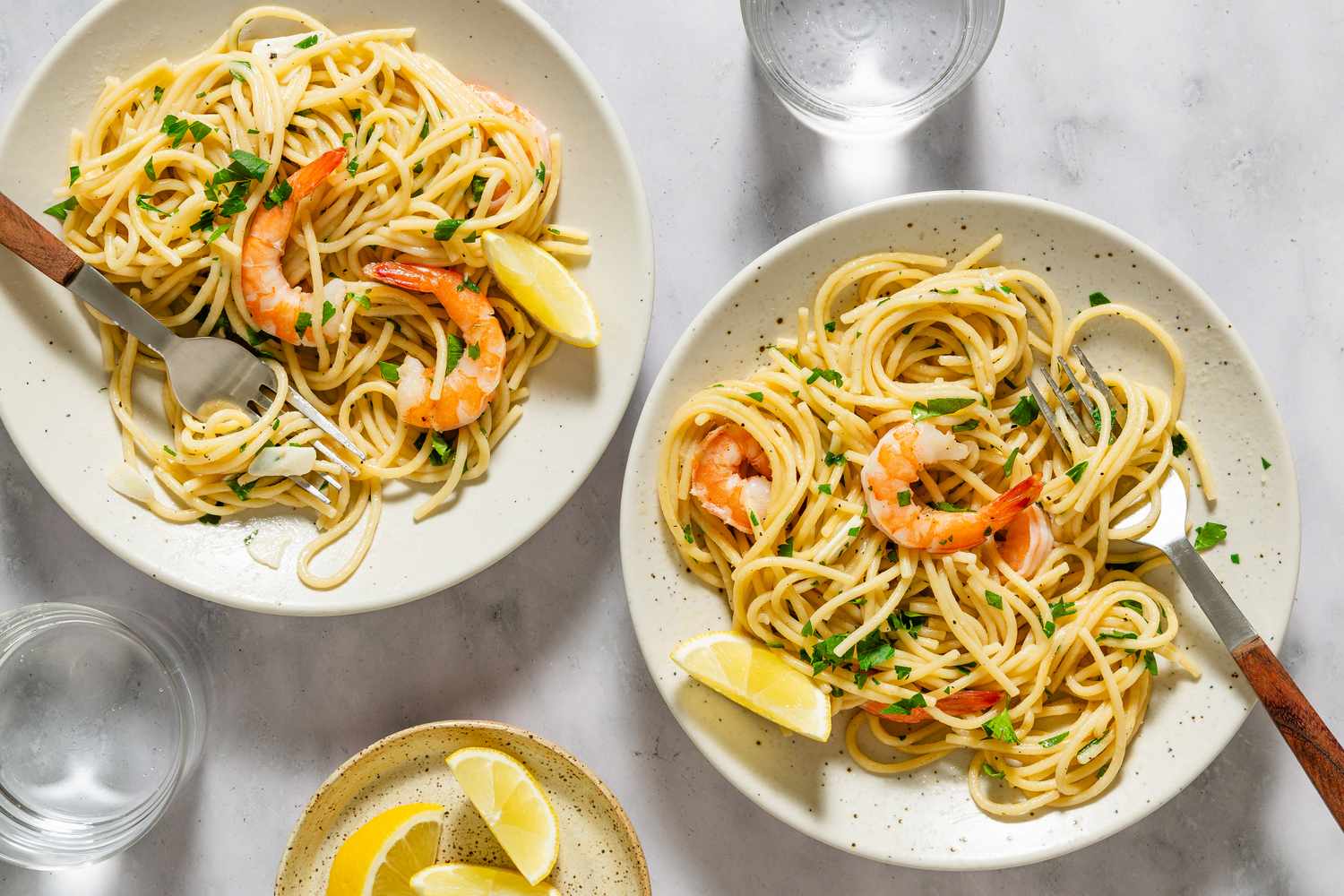 Two plates of shrimp pasta garnished with parsley and lemon wedges served with water glasses and a small bowl of lemon slices on the side
