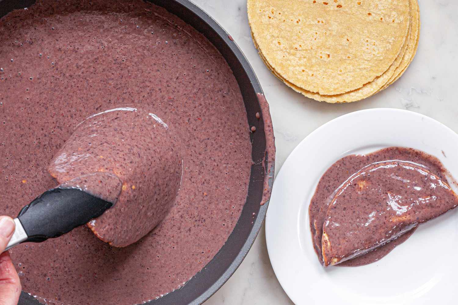 Corn tortillas topped with black bean sauce to show How to Make Enfrijoladas.