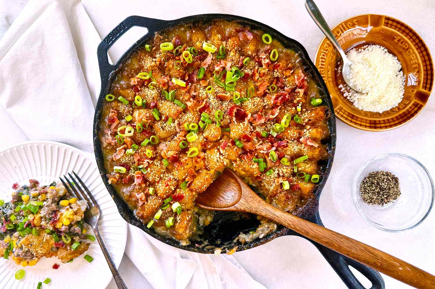 A castiron skillet containing a casserole with toppings such as green onions bacon and melted cheese served alongside plates