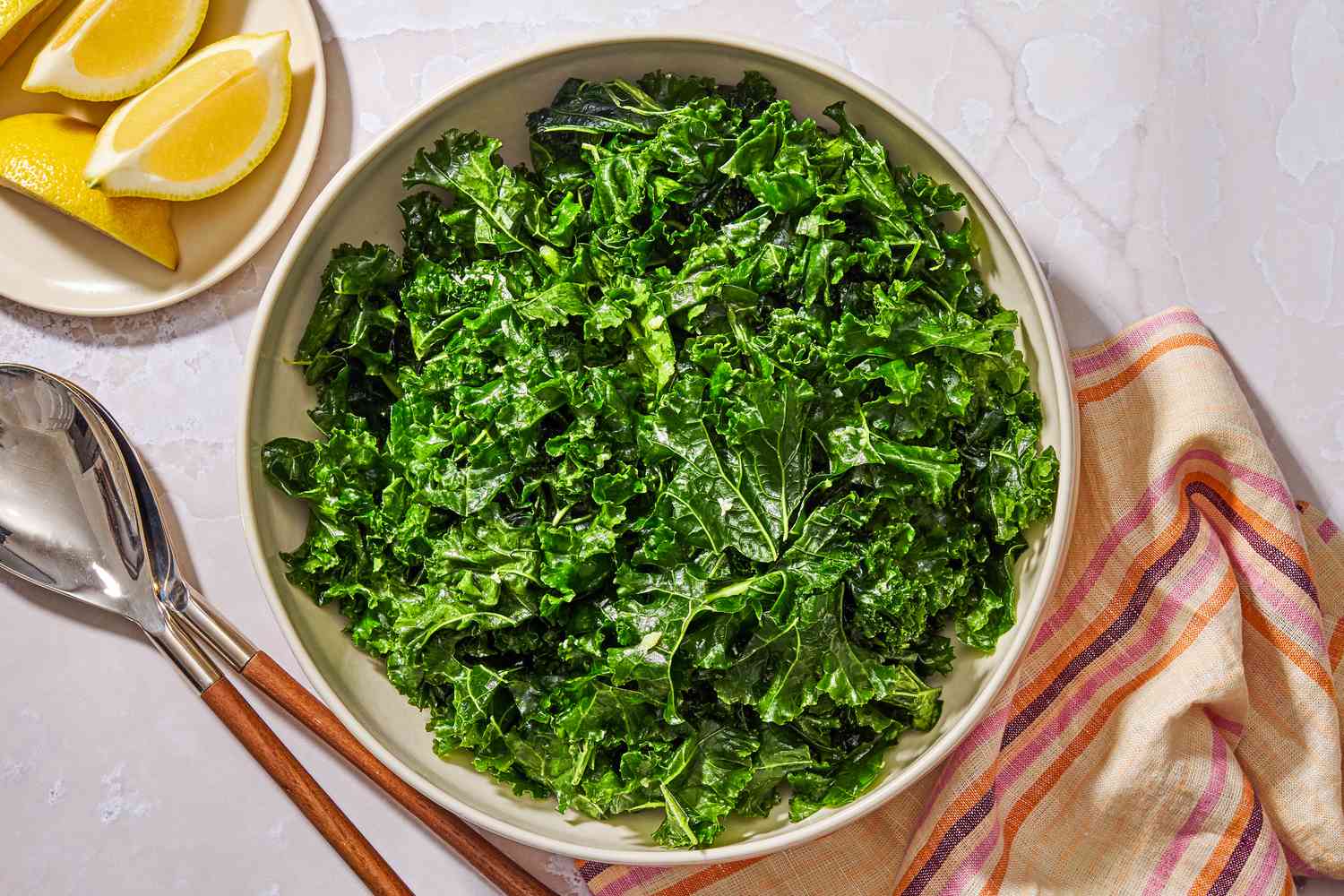 Overhead view of a large bowl of kale salad next to a plate of lemon slices, serving spoons and a cloth napkin