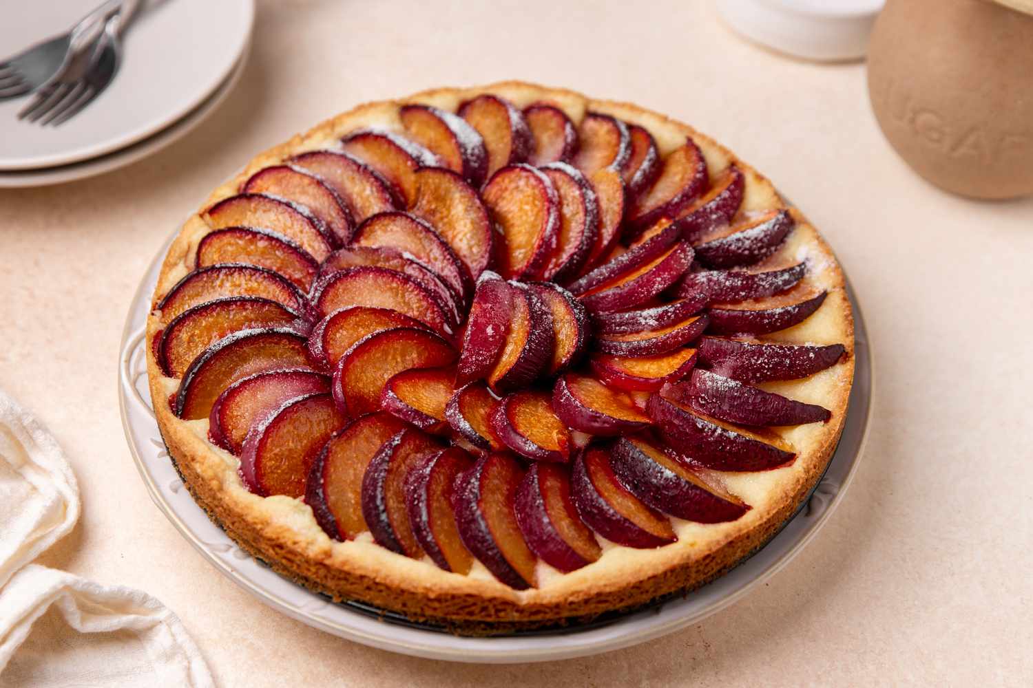 german plum tart at a table setting with a table linen, a stack of plates with some forks, and a vase