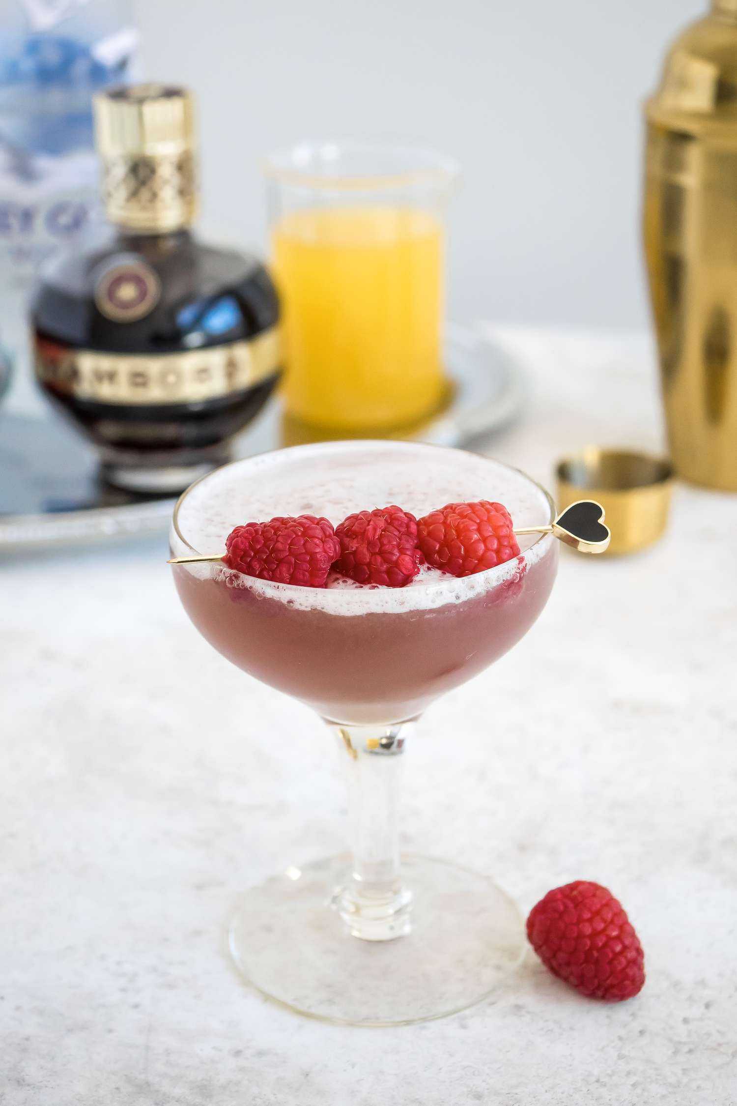 French Martini in a Glass Garnished with Raspberries and Surrounded by Shaker and a Tray with a Bowl of Raspberries, Vodka, Chambord, and Pineapple Juice 