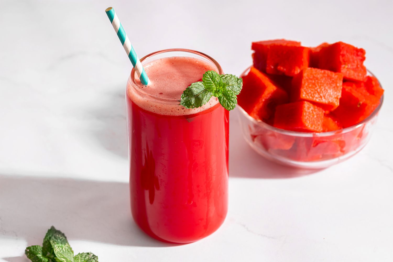 A glass of watermelon batido with a striped straw and fresh mint next to a bowl of watermelon pieces
