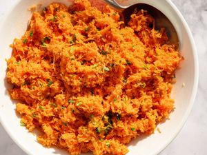 Shredded carrot salad in a white bowl with a serving spoon on a marble surface