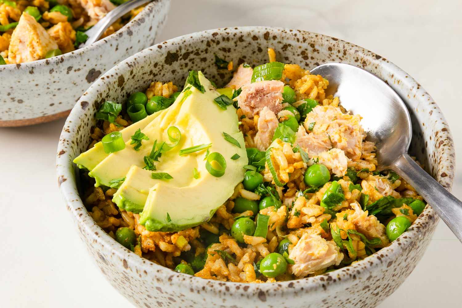 A bowl of rice mixed with peas salmon and topped with avocado slices served with a spoon