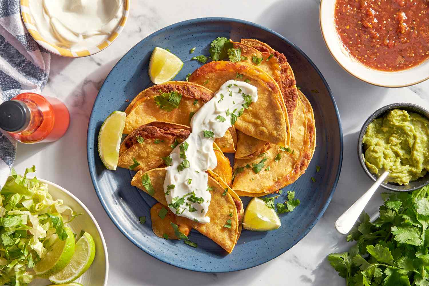 Bowl of cheesy baked tacos on a plate topped with sour cream and cilantro at a table setting with a bowl of lime wedges and shredded lettuce, bowl of sour cream, bowl of salsa, bowl of salsa, and a bunch of cilantro