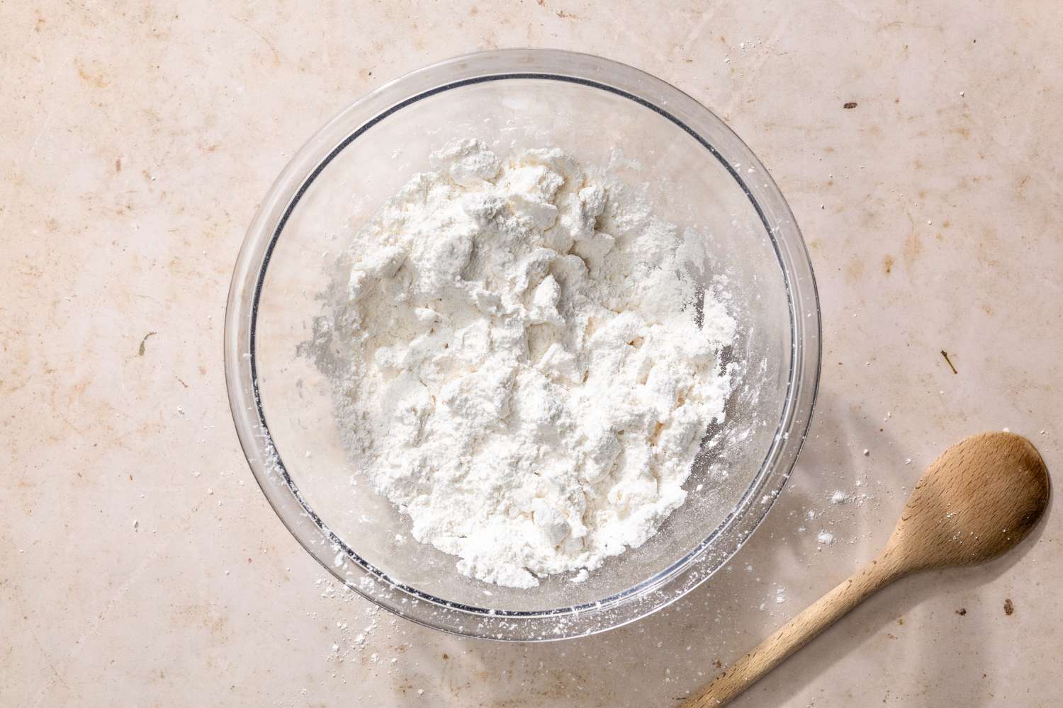 Overhead view of a clear glass bowl of the dry ingredients next to a wooden spoon for Mashed Potato Biscuits recipe