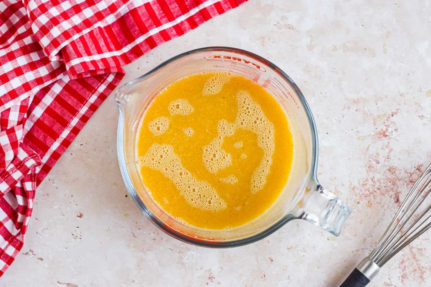 Wet Ingredients Whisked Together in a Pyrex Measuring Cup Next to a Red and White Checkered Kitchen Towel for Corn Dog Muffins Recipe