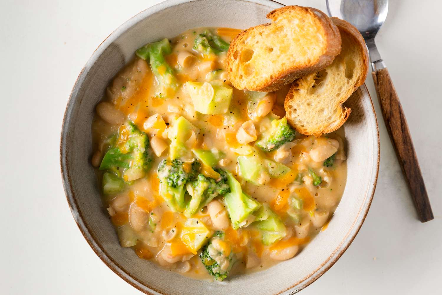 Overhead view of a stoneware bowl of Broccoli Cheddar Creamy Beans with two pieces of bread next to a spoon and all on a white background