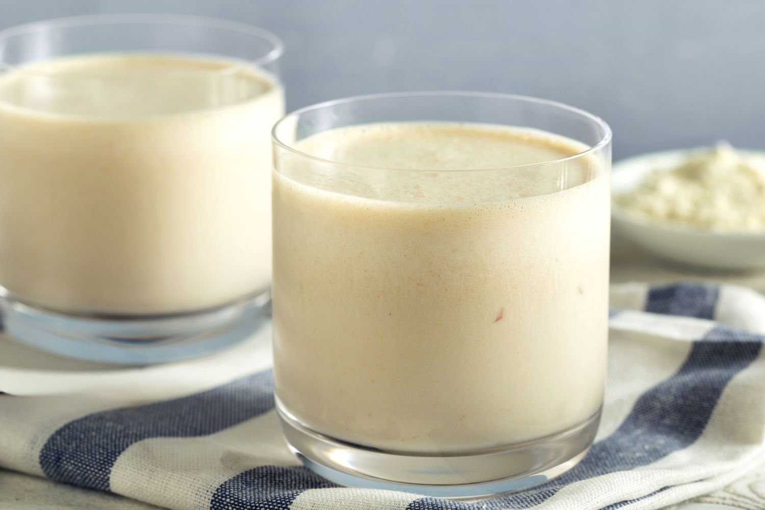 Two glasses of a creamy drink on a striped cloth protein powder in the background