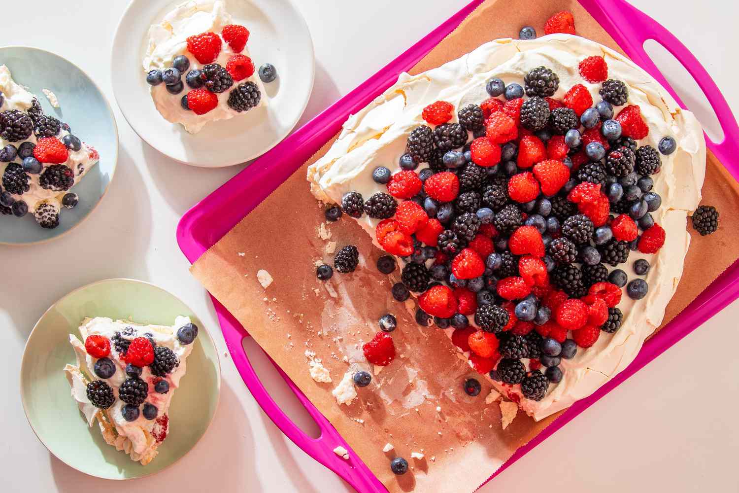 berry pavlova (with slices cut out) on a parchment paper lined tray next to three plates with slices of pavlova (overhead shot) 