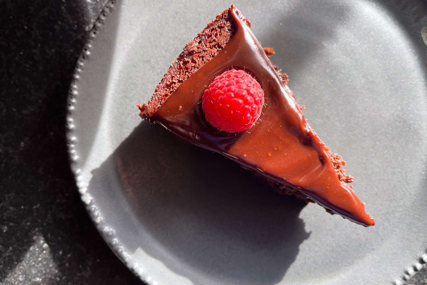 Slice of chocolate cake with glossy icing topped with a raspberry on a plate