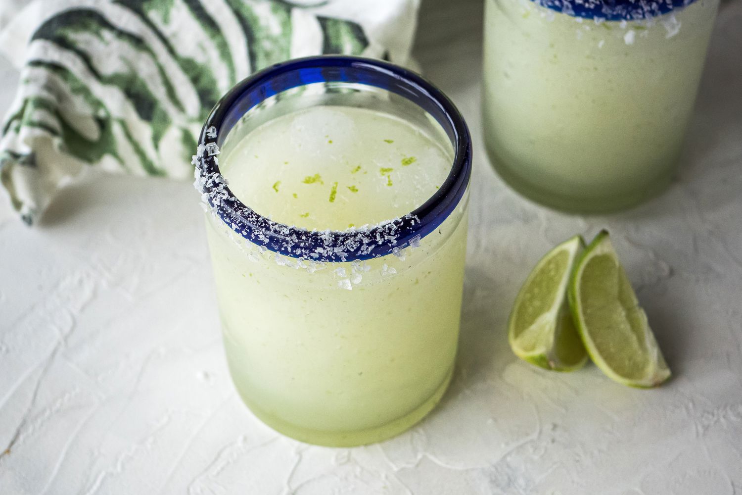 Best frozen margarita in a blue rimmed glass with limes in front and another glass and linen behind it.