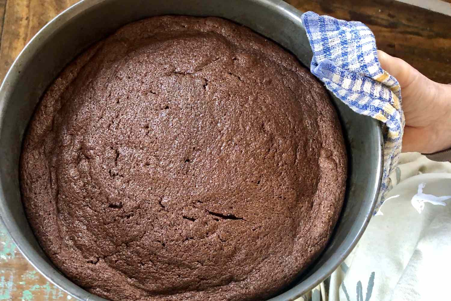 A person holding a freshly baked chocolate cake in a round pan with a cloth handle