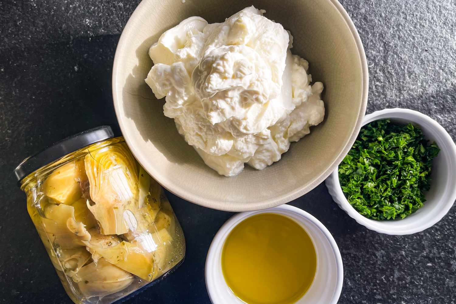Ingredients including a jar of preserved artichoke hearts a bowl of creamy cheese chopped herbs and olive oil in a bowl