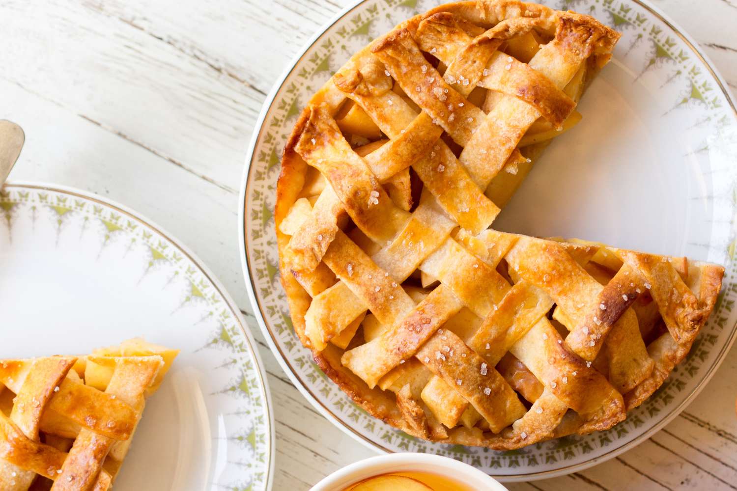 Overhead view of a plate with an apple pie with a slice removed next to another plate with a slice all on a white wooden tabletop