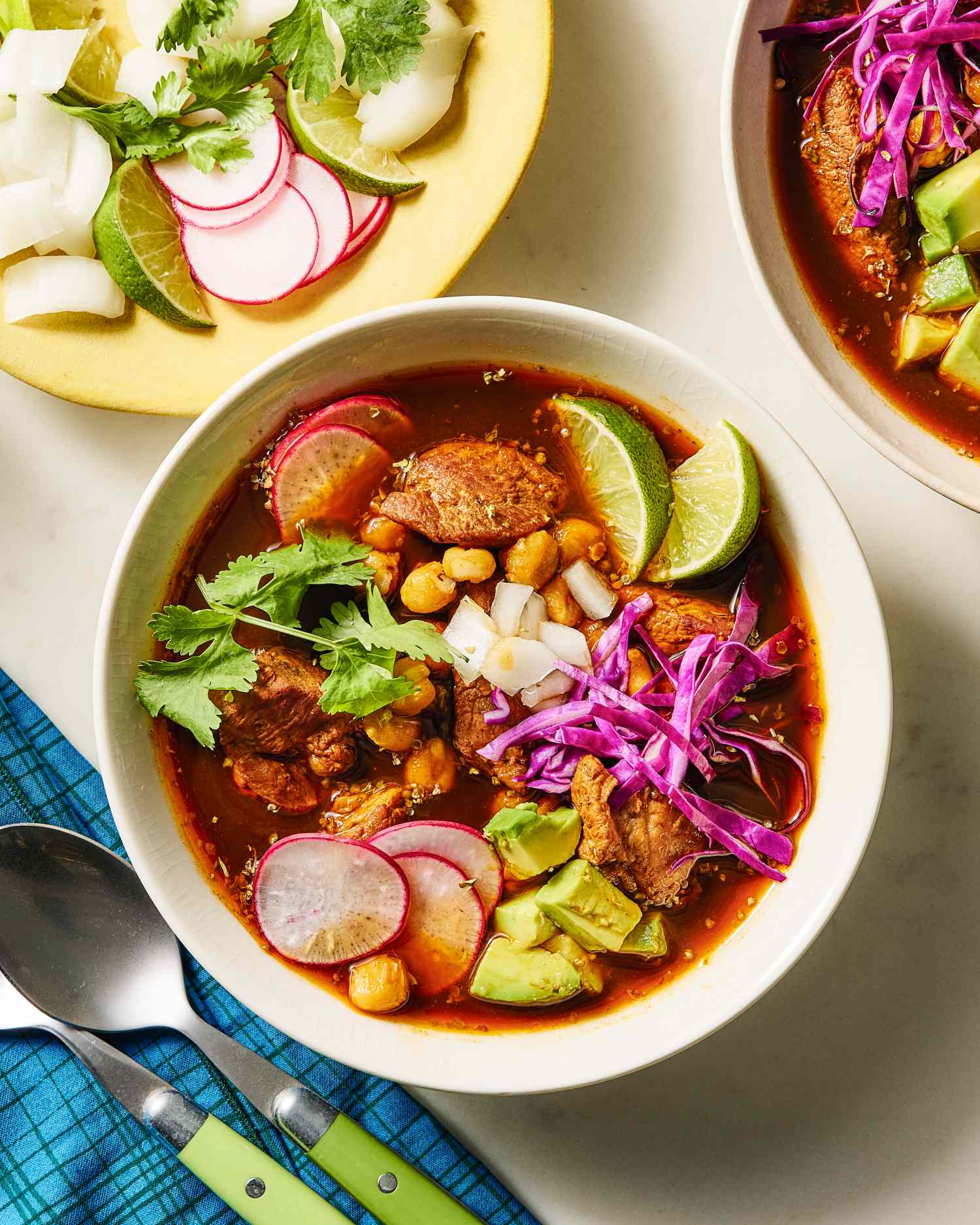 Bowl of pozole topped with radishes lime wedges cabbage and avocado accompanied by a plate with garnishes and a napkin with utensils