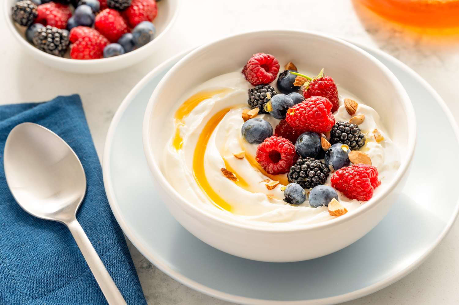 A bowl of yogurt topped with raspberries blackberries blueberries and honey a spoon and a cloth napkin are beside it