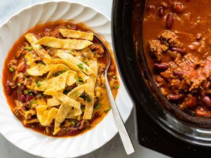 A bowl of beef and bean chili that's garnished with fried tortilla strips and sitting next to a slow cooker
