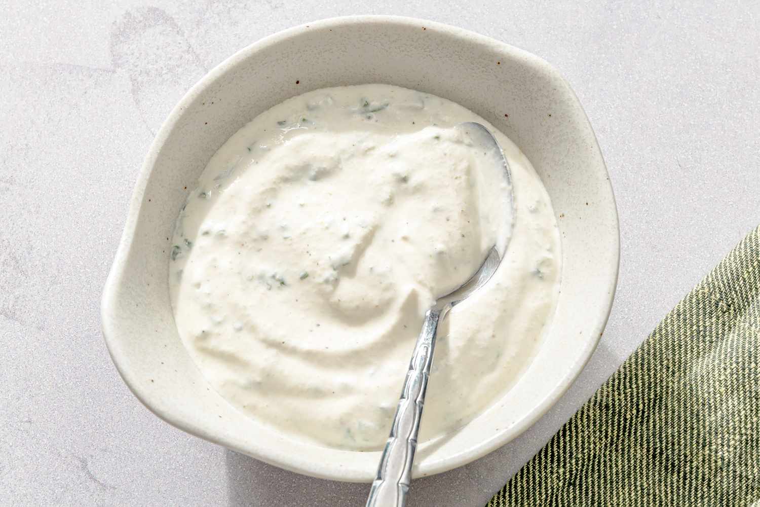 A bowl of sour cream and onion dressing with a spoon inside placed on a light surface next to a green cloth