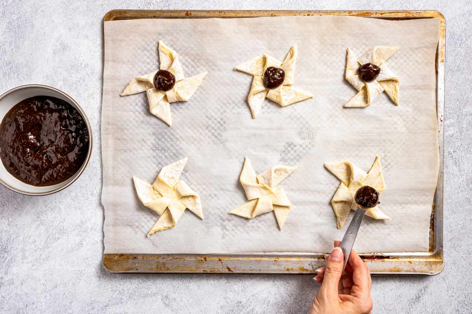 Preparing the Joulutortut (Finnish Christmas pastry) recipe with jam filling on a baking sheet a hand using a tool for shaping bowl of filling nearby